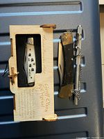 Photo showing two folding pocket knives and one small steel sharpener on a ribbed surface. One knife is in a wooden display box marked 'Geoff Olson Collection,' the other knife is used with dark handle and bolsters. Steel sharpener is compact with texture and chain attachment.