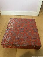 Photo of a rectangular red silk-covered box with gold traditional Chinese patterns, including pagodas and trees.