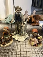 Three ceramic figurines displayed on fabric surface, showing an old man standing with a white goat and two seated girls in traditional clothing, one with a green dress and the other with a red headscarf and chicks.