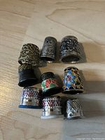 Top-down view of nine thimbles arranged on a wooden surface showing variety in colors, metal finishes, engravings and enamel floral decorations.