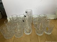 Fourteen crystal drinking glasses including two tall glasses with stickers and twelve shorter glasses with starburst and geometric cut designs on a wooden floor.