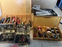 Four small tackle boxes filled with various fishing lures, fly fishing flies, spools of fishing line, hooks, weights, and metal tools arranged on a surface.
