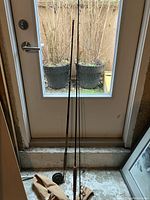 Two fly fishing rods with cork handles; one rod has an attached reel. A tan soft case is placed next to the rods. The items are displayed vertically against a glass door.