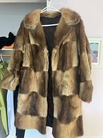 Front view of brown patchwork fur coat hanging on hanger against white wall. Patchwork of different shades of brown fur visible, with large fur collar and cuffs.