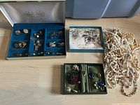 Wide view of three groups of costume jewelry including jewelry boxes with earrings, rings, brooches, cufflinks and loose white bead necklaces.