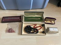 Photo of costume jewelry and pens arranged on floor including necklaces, earrings in boxes and plastic bag, two pens in original boxes, and a decorative ceramic box.