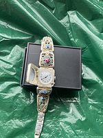 Close-up photo of a silver-tone women's wristwatch with a hinged cover and floral enamel accents on the band, placed on a dark box against a green background.