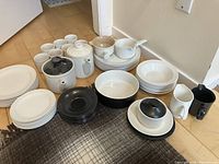 Full set of white and black ceramic dining pieces arranged on hardwood floor with a rug. Includes plates, cups, bowls, teapot, coffee pot, cream jug, gravy boat, and black accessories.