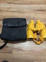 Black fabric carrying case with leather flap and yellow binoculars with strap, placed side by side on wooden floor.