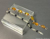 Two bracelets displayed on a clear acrylic stand, showing one with five oval amber-colored faceted stones and the other with black beads.