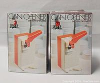Two vintage Black & Decker battery operated can openers new in original boxes showing front design with orange lever handles and white bases.