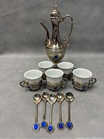 Full set showing the teapot at the back, five ceramic cups in metal holders, and five matching spoons in front. All pieces feature blue stone decorations and intricate metal embossing.
