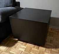 Black cube side table placed next to a sofa on wood parquet flooring, showing the tabletop and sides clearly.