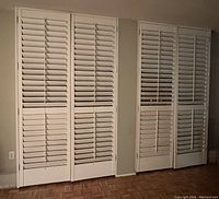 Four white painted custom California shutters consisting of three sets of two large one-piece panels and one set of two bi-fold panels shown against a wall.