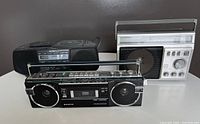 Three portable radios and players displayed side by side on a white surface, with Sanyo in front, Sony and Panasonic behind.