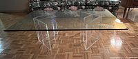 View of the glass coffee table showing the full tabletop and two clear acrylic base pieces from the front side in a living room setting.