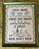 Photo showing framed cross-stitch piece with phrase and decorative embroidery in green wooden frame, laid on parquet floor.