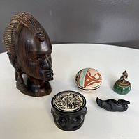 Photo of the full lot showing all five items on a white round table, highlighting the wooden head sculpture, ceramic orb, jade holder with brass lid and tongs, carved wooden trinket dish and carved black stone bird.