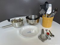 Full lot showing two stainless steel bowls, a Foley food mill, Corning Ware pie plate, mustard yellow crock with assorted kitchen utensils, citrus reamer, melon baller, and tea steeper arranged on a white round table.