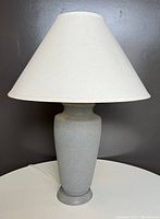 Matte grey ceramic ginger jar style table lamp with white fabric conical shade. Lamp base has a smooth, matte finish. Shade appears slightly aged or discolored.