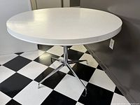 Front side view of the round white laminate dining table with chrome legs on a black and white checkered floor.