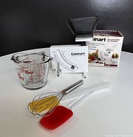 Cuisinart kitchen scale with original box, glass measuring cups, metal whisk and two silicone spatulas arranged on a surface