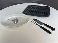 Four kitchen items displayed: 15-inch black broiler pan with removable drip tray on top, oval white ceramic serving platter underneath, metal dial-type meat thermometer on the platter, and a black-handled carving fork and knife set laid beside thermometer.