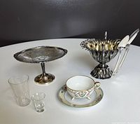 Lot showing all items together on white surface including silver-plated sugar bowl with spoons, pedestal tray, Nippon china tea cup and saucer, and two glass shot glasses.