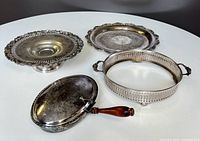 Four silver-plated serving accessories: silent butler with wooden handle, small tray, dish on pedestal, and decorative collar.