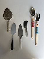 Seven assorted serving utensils arranged on white background showcasing various handles and blade types