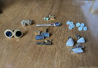 Overview of all cufflinks, tie clips, and button cuff stays displayed on wood surface.