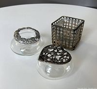 Clear glass square votive candle holder with dark metal grid frame, and two small round glass potpourri jars with decorative sculpted metal lids, one round lid with fruit/flower motif, one heart-shaped lid with floral openwork.
