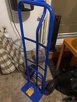 Blue steel hand truck dolly standing upright in garage on concrete floor, showing full frame including backrest, handles, wheels, and base plate.