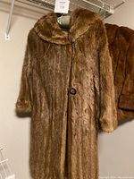 Full view of the long brown fur coat hanging, showing its length, collar shape, and two buttons visible in front.