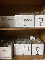 Shelving unit with multiple original boxes of Ikea Svalka wine glasses labeled by type (Red, White, Mix). Each box holds 6 glasses.