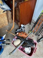 Full assortment view of sporting equipment showing bats, gloves, balls, hockey sticks, and skates in a plastic container and leaning against cabinet