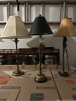 Photo showing all three tall buffet lamps with different colored shades and brass/bronze toned bases lined up on cardboard boxes.