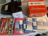 Shows the brown View-Master stereo viewer, a collection of View-Master slide packets with colorful covers, and the box for the light attachment.