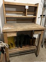 Photo of wood tool bench showing overall structure including drawers, upper shelves, and lower slatted shelf. Visible wear and several pairs of boots stored beneath.