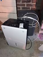 White Kenmore dehumidifier positioned in front of the darker GE unit on a concrete floor in a basement setting.