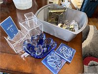 Lot overview showing blue glass bowl with glass beads, two blue decorative ceramic tiles, clear acrylic display stands, and plastic bin containing door knobs and vase.
