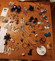 Multiple pairs and singles of earrings arranged on a wooden table, showing a variety of styles and materials including beaded, metal, and pearls