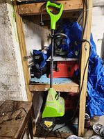 Full view of the green electric snow shovel with handle and power cord, placed indoors against shelving.