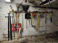 Photo showing various garden tools hanging on a wall including rakes, weed wacker, measuring tape, and Red Sox floor mats on ground