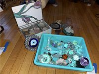 Photo showing stained glass jewelry/trinket box with a floral design, evil eye wall art, and a plastic bin containing various crystals and stones including amethyst.