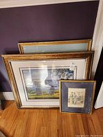 Three framed art pieces leaning against a wall: one large golden frame, one medium frame with a harbor scene, one smaller vintage fashion print