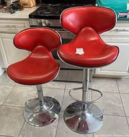 Two red adjustable bar stools with chrome pedestal bases standing on tiled floor in kitchen setting