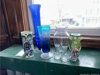 Seven vases lined up on a green surface near window, showing variety of colors and styles including green ceramic with floral motif, tall blue glass, and smaller clear glass vases.