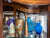 Photo showing five vases on glass shelving: ceramic blue-purple drip glaze, floral ceramic, hammered metal bronze, large blue glass, small electric blue glass with metal decoration