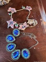 Top view of both necklaces showing vibrant pink and purple floral design on one and large blue faceted stones on the other.